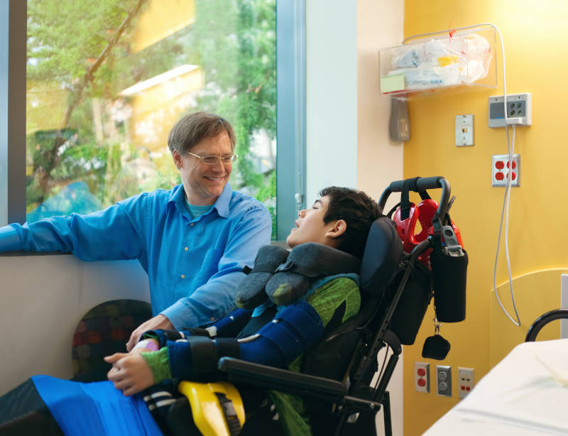 Smiling father sitting next to disabled son in wheelchair by  hospital bed,  talking  together